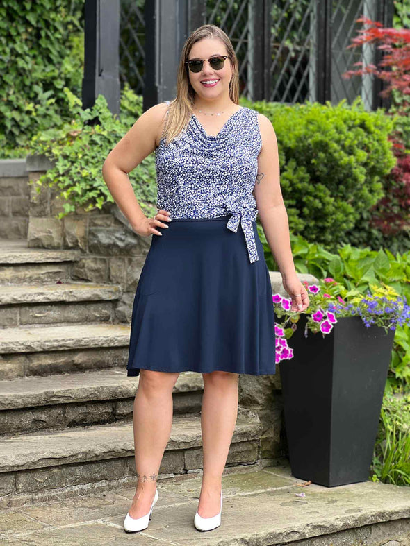 Miik model Christal (5'3", medium) smiling wearing Miik's Ada reversible draped cowl neck tank in baby's breath print along with a Blair belt in the same matching print and a flouncy skirt in navy