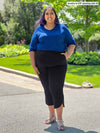 Miik model plus size Sureka (5'9", 3x) smiling with hand on pockets wearing Miik's Akira tulip hem capri pant in black along with a ink blue blouse tucked in #color_black