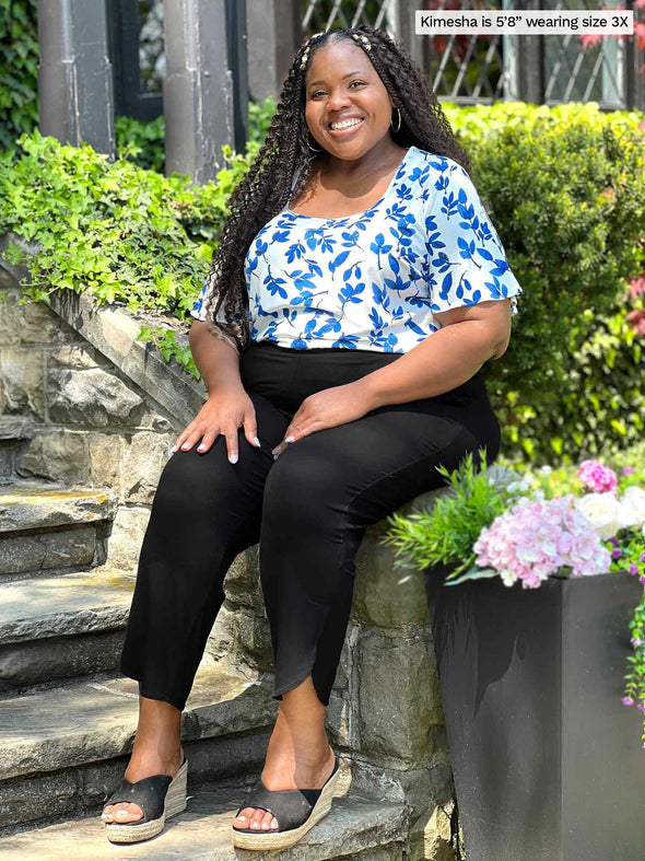 Miik model plus size Kimesha (5'8", 3x) sitting and smiling wearing Miik's Akira tulip hem capri pant in black with a top in ink leaf print #color_black