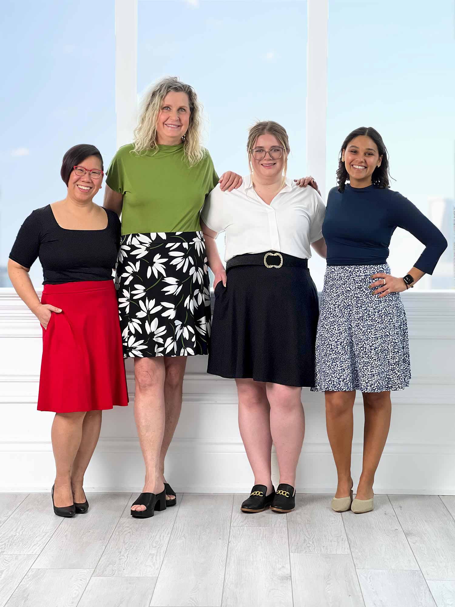 Miik models Mary-Ann, Carolyn, Bri and Meron standing next to each other, smiling, all wearing the new Miik skirt: the Alara pocket swing skirt. Colour available are: poppy red, white lily print, black, baby&