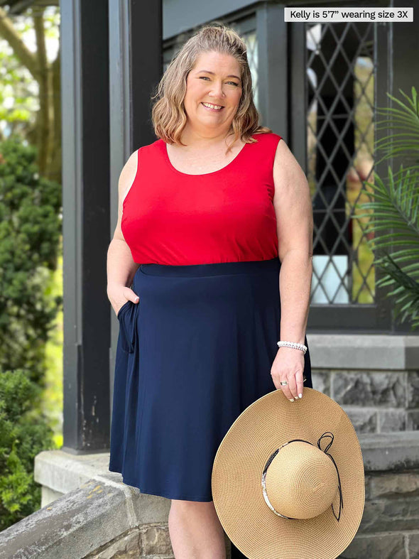 Miik model plus size Kelly (5'7", 3x) smiling with one hand on the pockets wearing a poppy red tank top along with Miik's Alara pocket swing skirt in navy while holding a beach hat