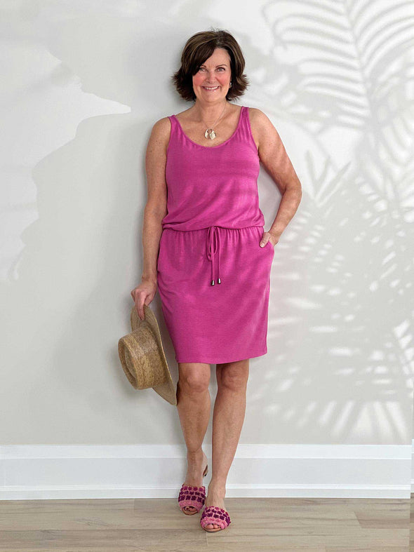 Miik founder Donna (5'6", small) smiling while standing in front of a white wall wearing Miik's Amy drawstring pocket dress in pink #color_pretty-in-pink