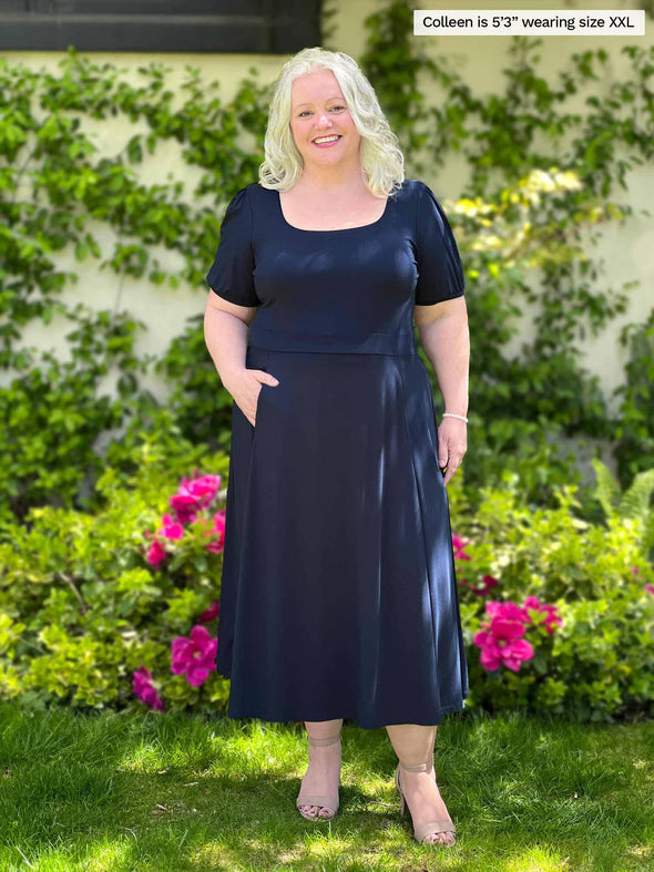 Woman standing in front of a garden wearing Miik's Anika puff sleeve midi dress in navy. #color_navy