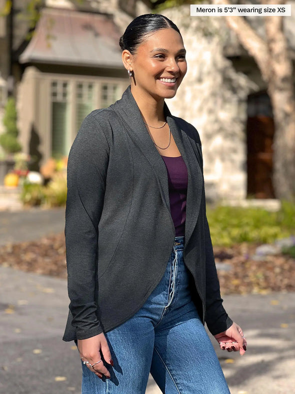 Miik model Meron (5'3", xsmall) smiling while standing sideway wearing Miik's Anne soft stretch blazer in charcoal with a port tank and jeans #color_charcoal