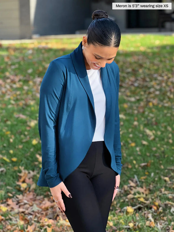 Miik model Meron (5'3", xsmall) smiling and looking down wearing Miik's Anne soft stretch blazer in teal with a white tank and a black dress pant #color_teal