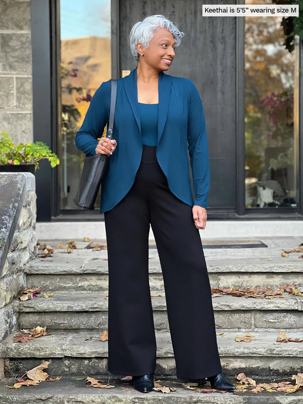 Miik model Keethai (5'5", medium) looking away while standing in front of a door entry wearing a wide leg pant in black with Miik's Anne soft stretch blazer in teal and a matching colour tank #color_teal 