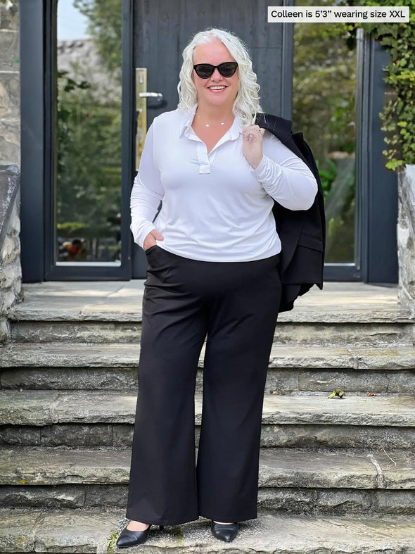 Miik model Colleen (5'3", xxl) smiling wearing Miik's Arianna collared shirt in white with a black wide leg pant and a blazer in the same colour over her shoulders #color_white 