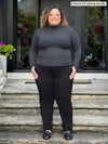 Miik model Carley (5'2", xxlarge) smiling wearing a turtleneck top in charcoal along with Miik's Ashton patch pocket jogger in black #color_black