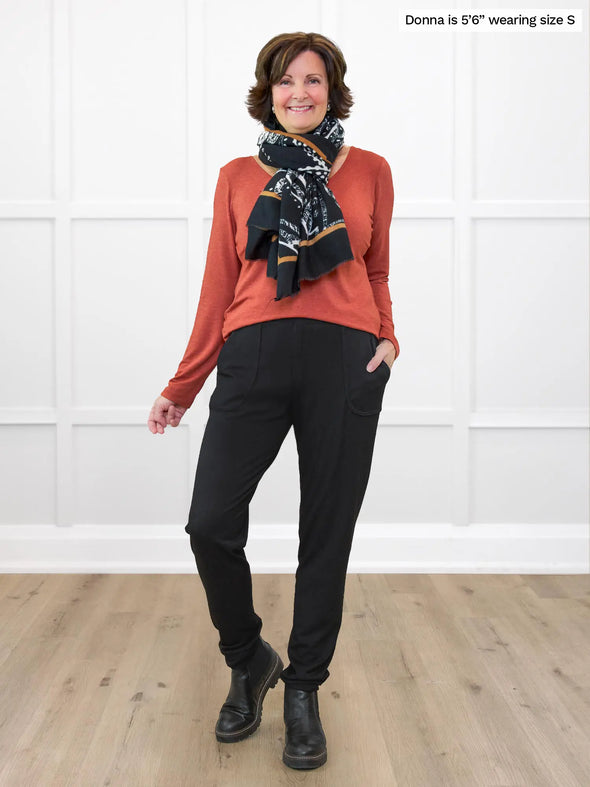 Miik founder Donna (5'6", small) smiling while standing in front of a white wall wearing Miik's Ashton patch pocket jogger in black with a paprike melange long sleeve top and scarf #color_black