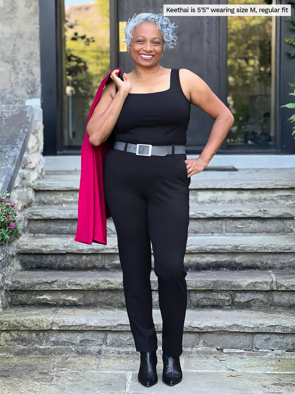 Miik model Keethai (5'5", medium) smiling wearing Miik's Asia mid-rise slim pant in black, regular fit, with a belt and a tank top in the same colour #color_black