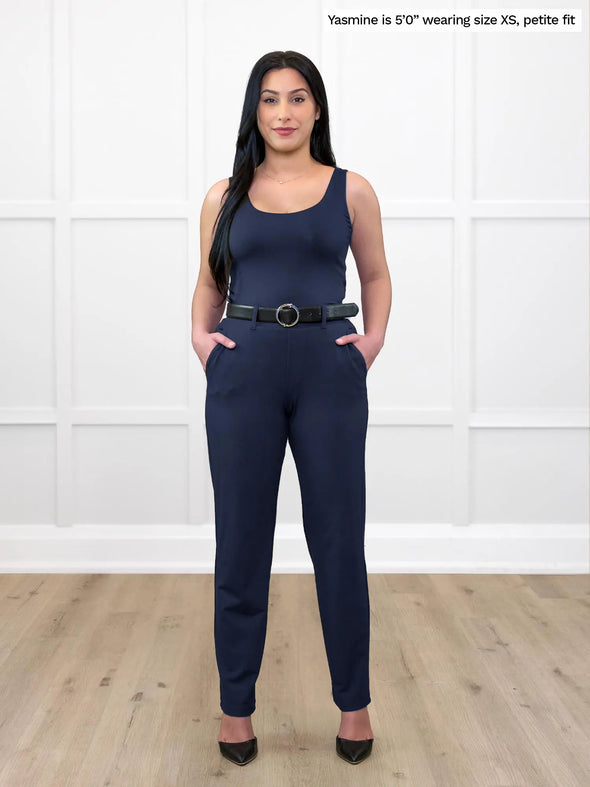 Miik model Yasmine (5'0", xsmall, petite) standing in front of a white wall wearing Miik's Asia mid-rise slim pant in navy, petite fit, with a belt and a square neck tank top #color_navy