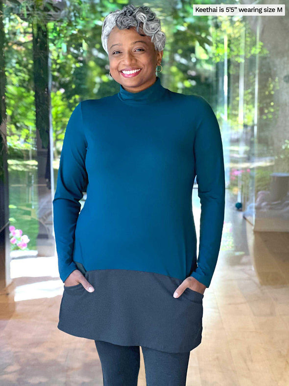 Woman standing in front of a window wearing Miik's Brooklin mock neck pocket tunic in teal and charcoal with leggings.