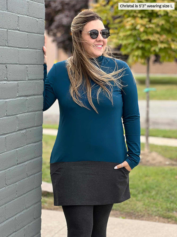 Woman standing next to a building wearing Miik's Brooklin mock neck pocket tunic in teal and charcoal with charcoal leggings