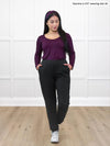 Miik model Yasmine (5'0, xsmall. petite) standing in front of a white wall looking down wearing Miik's Cameron luxe fleece jogger with pockets in black with a long sleeve top in port #color_black