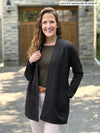 Miik model Lex (5'2", xsmall) smiling wearing Miik's Carmen luxe fleece cardigan with pockets in black, petite fit, along with a olive tee and a white jeans #color_black