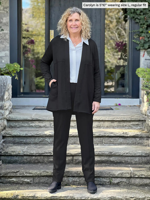 Miik model Carolyn (5'10", large) smiling wearing Miik's Carmen luxe fleece cardigan with pockets in black, regular fit, with a dress pant in the same colour and a collared shirt in white #color_black