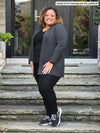 Miik model Carley (5'2", xxlarge, petite) smiling wearing an all black outfit along with Miik's Carmen luxe fleece cardigan with pockets in charcoal, petite fit, and sneakers #color_charcoal