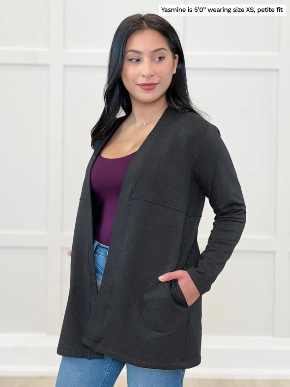 Miik model Yasmine (5'0", xsmall, petite) looking down wearing Miik's Carmen luxe fleece cardigan with pockets in charcoal, petite fit, with a port tank top and jeans #color_charcoal