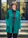 Miik model Jenna (5'9", small) smiling wearing Miik's Carmen luxe fleece cardigan with pockets in jade melange, regular fit, with a turtleneck top in charcoal and black leggings #color_jade-melange