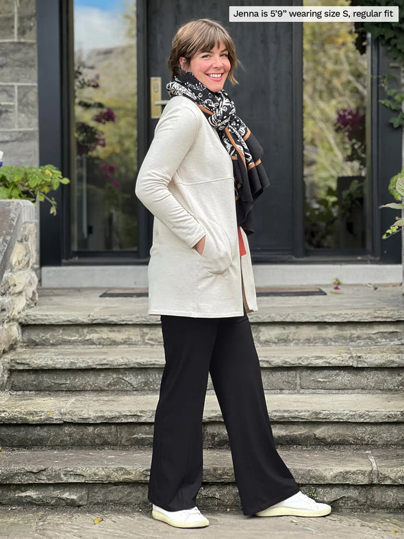 Miik model Jenna (5'9", small) smiling while standing sideway 
wearing a black pant along with a paprika melange tee and Miik's Carmen luxe fleece cardigan with pockets in oatmeal melange, regular fit, and a scarf #color_oatmeal-melange