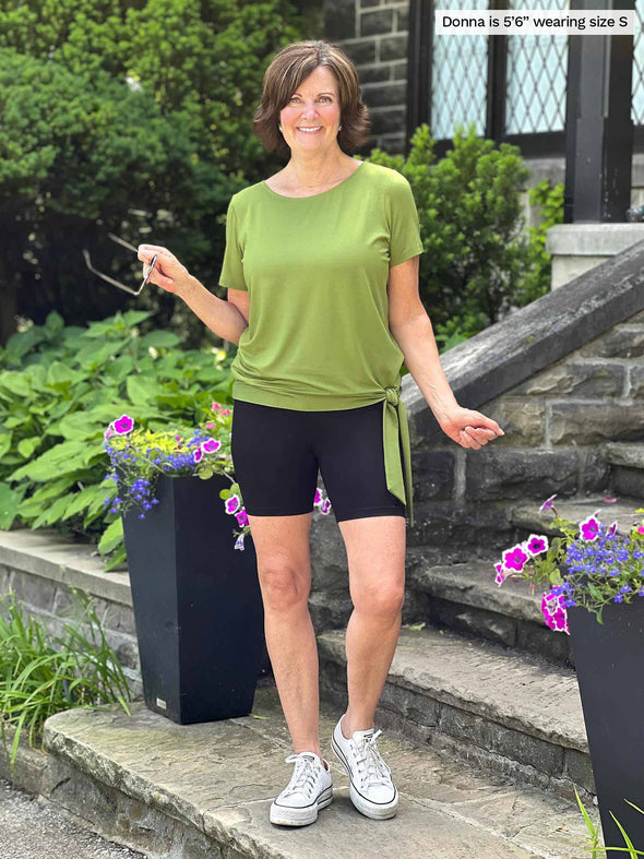 Miik founder Donna (5'6", small) smiling wearing Miik's Chalha high waisted mid thigh biker short in black with a green moss tie top and sneakers 