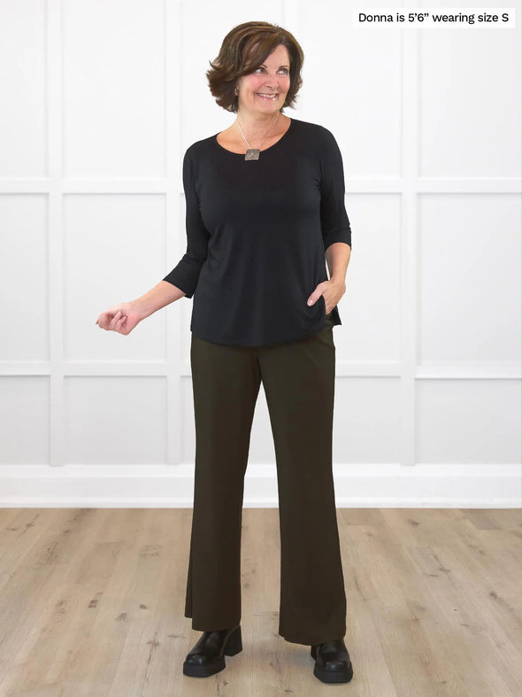 Miik founder Donna (5'6", small) smiling while looking away in front of a white wall studio wearing a olive high waisted straight leg pant and Miik's Chris 3/4 sleeve scoop neck top in black and boots 
#color_olive 