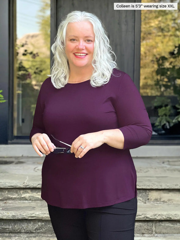 Miik model Colleen (5'3", xxlarge) smiling wearing Miik's Chris 3/4 sleeve scoop neck top in port with a dress pant in black while holding a sunglass 
#color_port 