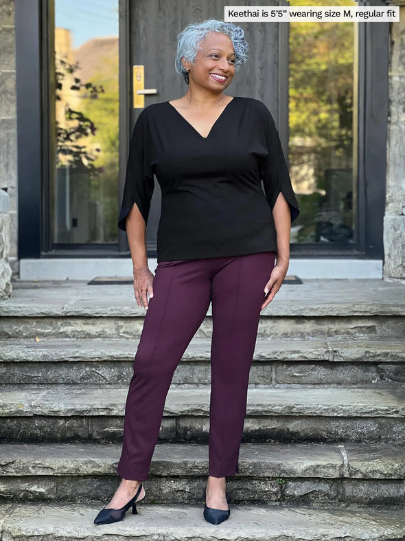 Miik model Keethai (5'5", medium) smiling and looking away wearing Miik's Christal pull-on pintuck ankle pant in port, regular fit, along with a v-neck blouse in black and heels 
#color_port 