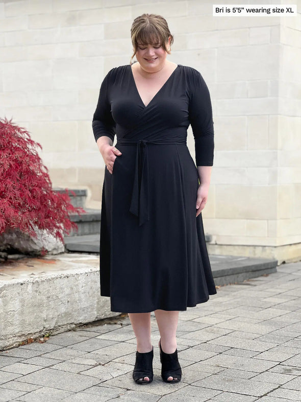 Miik model Bri (5'5", xlarge) smiling and looking down wearing Miik's Claudine tie waist midi dress in black and matching colour high heels #color_black