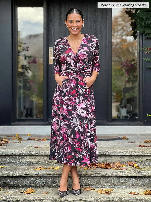 Miik model Meron (5'5", xsmall) smiling wearing Miik's Claudine tie waist midi dress in midnight floral with both hands on pockets #color_midnight-floral