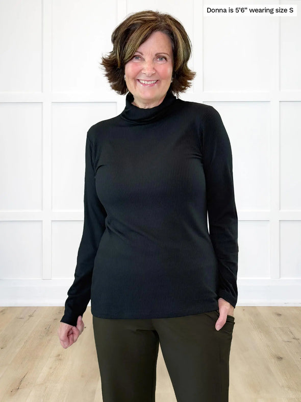 Miik founder Donna (5'6", small) smiling while standing in front of a white wall wearing Miik's Cora ribbed turtleneck top in black with a olive pant #color_black