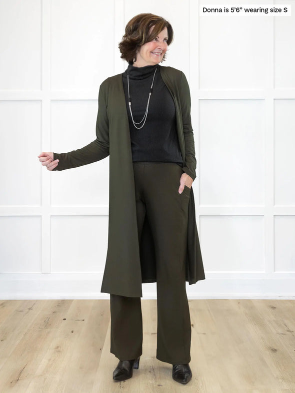 Miik founder Donna (5'6", small) smiling and looking away wearing Miik's Cora ribbed turtleneck top in black along with a duster cardigan and wide leg pant in the same olive colour #color_black