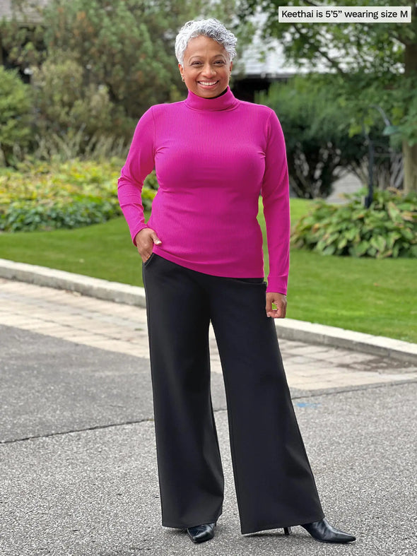 Miik model Keethai (5'5", medium) smiling wearing Miik's Cora ribbed turtleneck top in bright pink with black pants #color_bright-pink