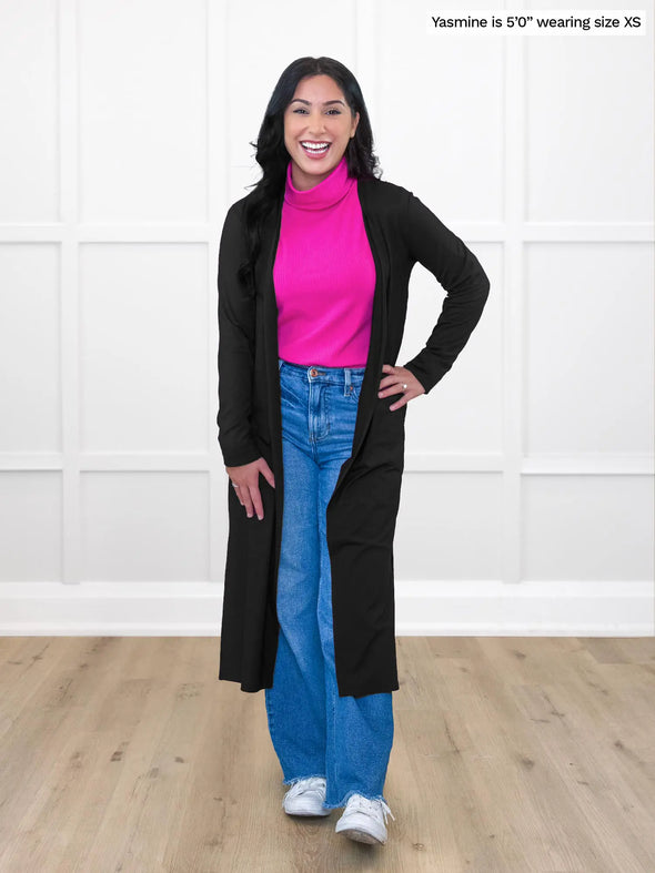 Miik model Yasmine (5'0", xsmall, petite) smiling wearing Miik's Cora ribbed turtleneck top in bright pink along with jeans and a duster cardigan in black #color_bright-pink