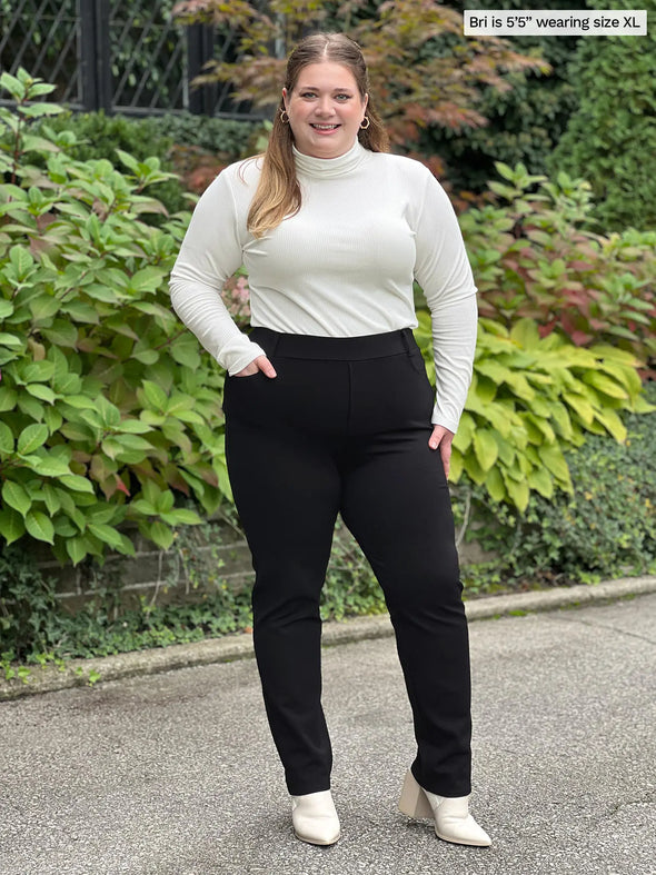 Miik model Bri (5'5", xlarge) smiling wearing a dress pant in black with Miik's Cora ribbed turtleneck top in natural with matching boots #color_natural