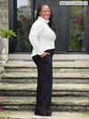 Miik model Marilyn (5'2", medium) standing side way and smiling wearing Miik's Cora ribbed turtleneck top in natural with a black wide leg pant #color_natural