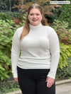 Miik model Bri (55", xlarge) smiling wearing Miik's Cora ribbed turtleneck top in natural with a black pant #color_natural 