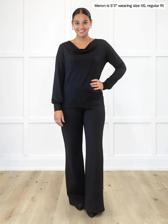 Miik model Meron (5'3", xsmall) smiling while standing in front of a white wall wearing Miik's  Devina draped cowl neck long sleeve top in black, regular fit, untucked with a wide straight leg pant in the same colour #color_black