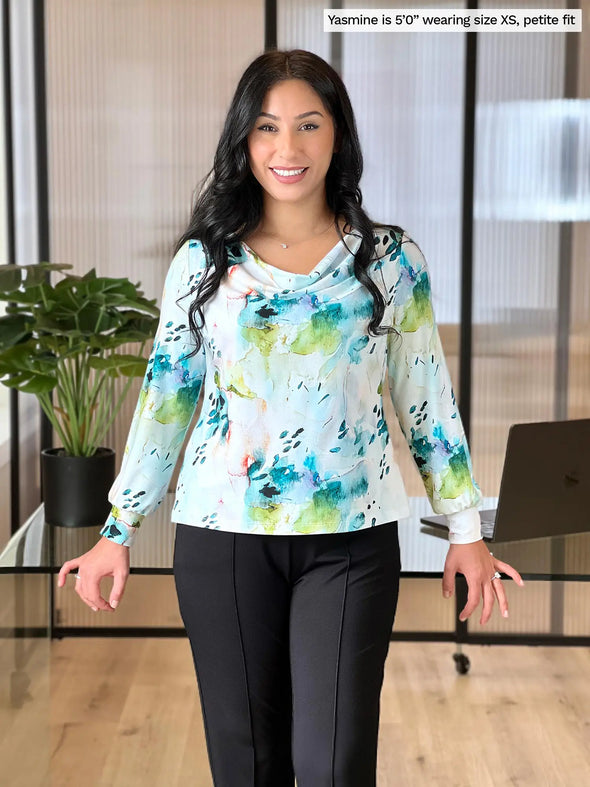 Miik model Yasmine (5'0", xsmall, petite) smiling while leaning against to a office desk wearing Miik's  Devina draped cowl neck long sleeve top in water lily, petite fit, with a black dress pant #color_water-lily