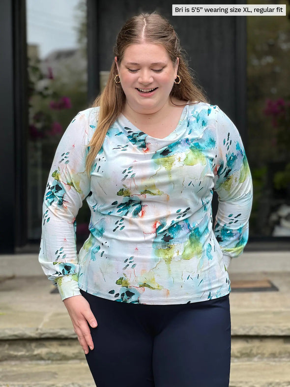 Miik model Bri (5'5", xlarge) smiling and looking down wearing Miik's  Devina draped cowl neck long sleeve top in water lily, regular fit, with a navy pant #color_water-lily