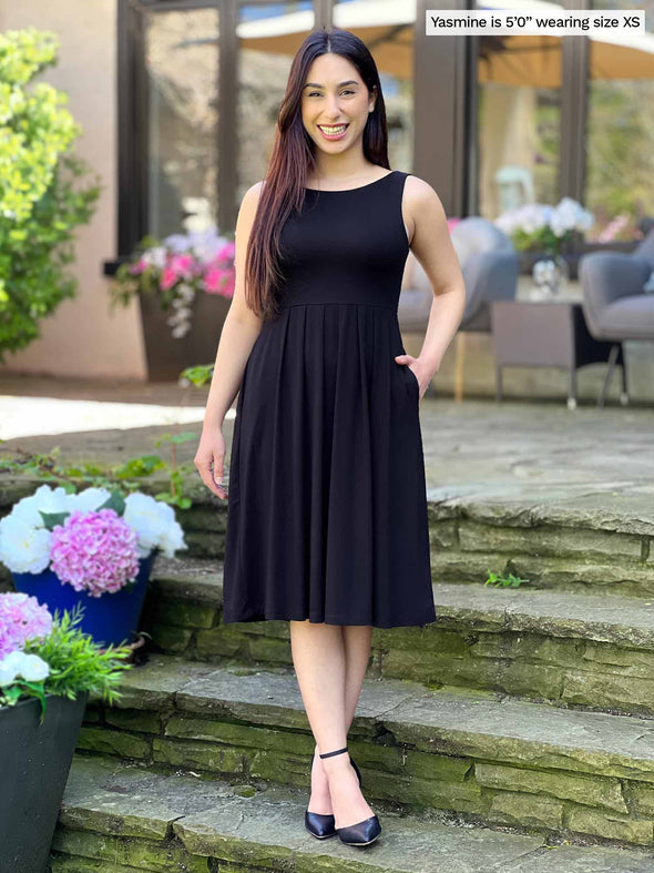 Miik model Yasmine (5'0", xsmall, petite) smiling wearing Miik's Ela reversible pleated sleeveless pocket dress in black 
