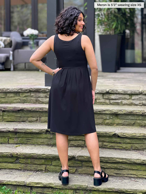 Miik model Meron (5’3”, xsmall) standing with her back towards the camera showing the back of Miik's Ela reversible pleated sleeveless pocket dress in black