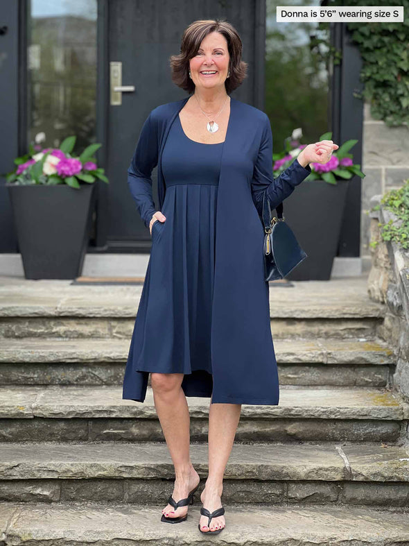 Miik founder Donna (5'6", small) smiling while standing on a stairway wearing Miik's Ela reversible pleated sleeveless pocket dress in navy along with a duster in the same colour 
