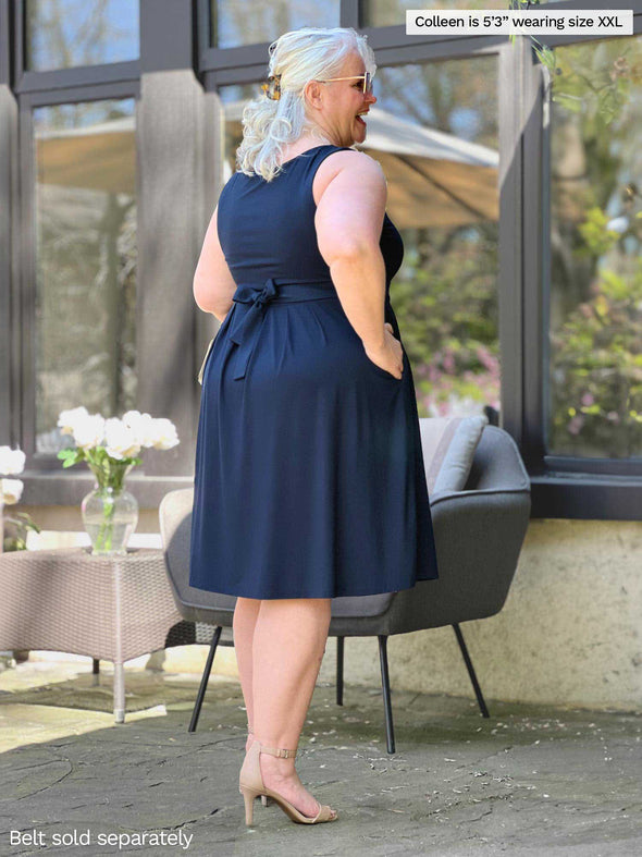 Miik model Colleen (5'3", xxlarge) standing with her back towards the camera showing the back of Miik's Ela reversible pleated sleeveless pocket dress in navy with a Blair belt in the same colour 