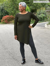 Miik model Keethai (5'5", medium) smiling and looking away wearing a charcoal legging with Miik's Elsie asymmetrical long sleeve tunic in olive #color_olive 