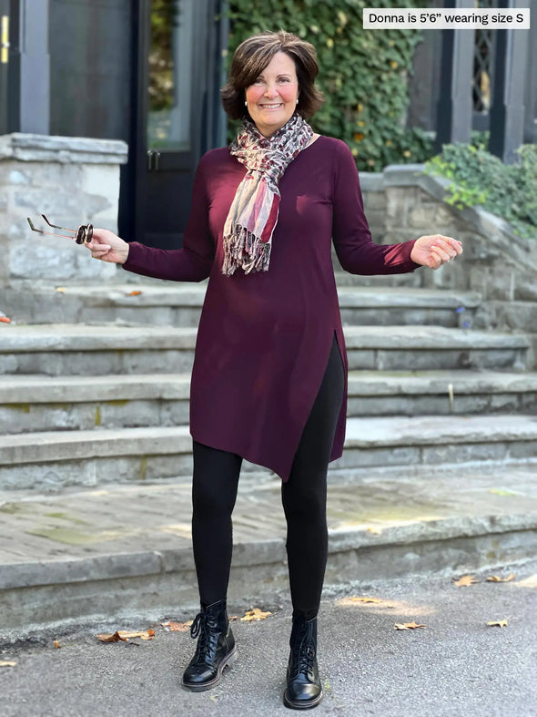 Miik founder Donna (5'6", small) smiling with her arms open wearing a black legging and boots along with Miik's Elsie asymmetrical long sleeve tunic in port and a scarf in animal print #color_port