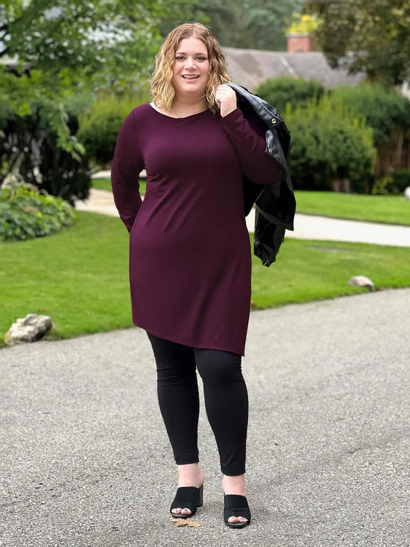 Miik model Bri (5'5", xlarge) smiling wearing Miik's Elsie asymmetrical long sleeve tunic in port with a black legging and a leather jacket over her shoulders #color_port