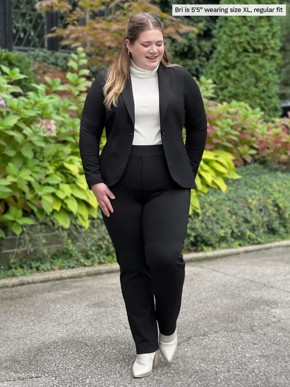 Miik model Bri (5'5", xlarge) smiling and looking down wearing Miik's Emily soft blazer in black, regular fit, with a dress pant in the same colour and a turtleneck top in natural and boots #color_black