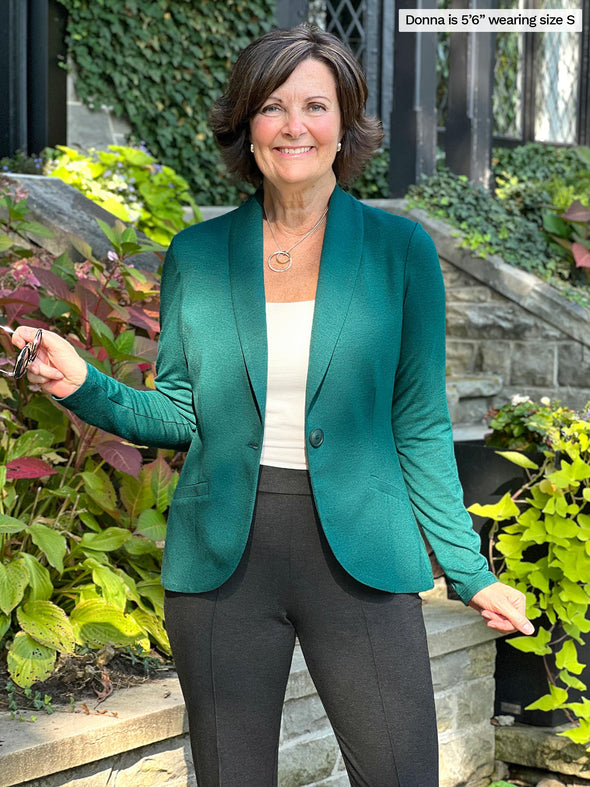 Woman standing in front of house wearing Miik's Emily soft blazer in green jade melange over a white top and pants.
