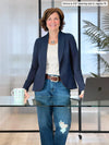 Miik founder Donna (5'6", small) smiling while standing behind a office desk wearing Miik's Emily soft blazer in navy, regular fit, with a natural ribbed tee, jeans and a belt #color_navy